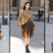 A imagem mostra três personalidades em looks de street style durante um evento de moda. À esquerda, um homem usa conjunto oversized de jeans escuro, sem camisa por baixo, carregando uma maleta de madeira. No centro, uma mulher veste blusa dourada de tricô com saia assimétrica de estampa xadrez, combinada com bota preta e bolsa estruturada. À direita, outra mulher aparece com um vestido longo off-white ajustado, de mangas bufantes, completando o visual com óculos escuros e bolsa preta