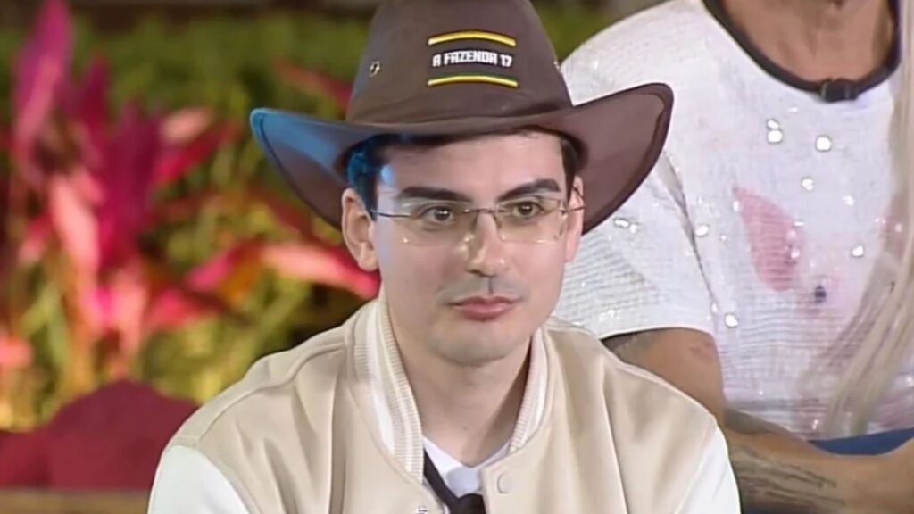 Dudu Camargo aparece sentado, usando um chapéu marrom com o logotipo do programa “A Fazenda 17”, óculos de grau e jaqueta bege com mangas claras. Ele está em um ambiente decorado com plantas e almofadas coloridas, participando de uma gravação do reality show. Ao fundo, vê-se parcialmente outra pessoa vestindo uma camiseta branca brilhante.