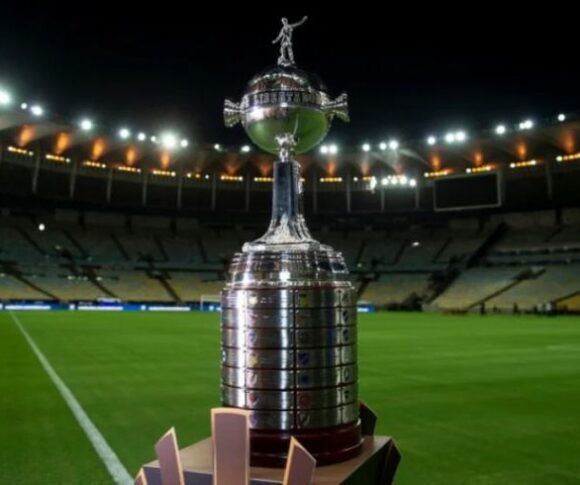 A imagem mostra o troféu da Copa Libertadores em destaque, posicionado em um estádio de futebol. O troféu é prateado, com uma base larga e arredondada, e no topo há uma pequena escultura de um jogador de futebol em ação. Ao fundo, vê-se um estádio bem iluminado, com arquibancadas vazias e um gramado verde em primeiro plano.
