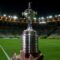 A imagem mostra o troféu da Copa Libertadores em destaque, posicionado em um estádio de futebol. O troféu é prateado, com uma base larga e arredondada, e no topo há uma pequena escultura de um jogador de futebol em ação. Ao fundo, vê-se um estádio bem iluminado, com arquibancadas vazias e um gramado verde em primeiro plano.