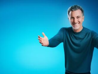 Luciano Huck aparece sorridente, com expressão amigável e convidativa. Ele tem cabelo grisalho, penteado para trás, barba bem aparada e está vestindo uma camisa de manga comprida azul. Sua postura, com os braços estendidos para frente, transmite entusiasmo e proximidade, reforçando uma imagem carismática e acolhedora diante do fundo azul vibrante