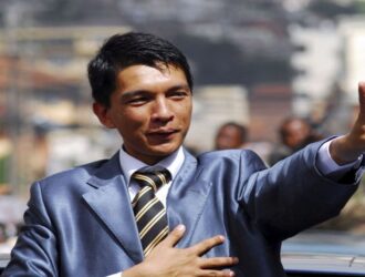 A imagem mostra Andry Rajoelina, presidente de Madagascar que fugiu após os protestos, em um momento público. Ele está de pé em um veículo de teto aberto, vestindo um terno azul brilhante, camisa branca e gravata listrada nas cores preto e dourado