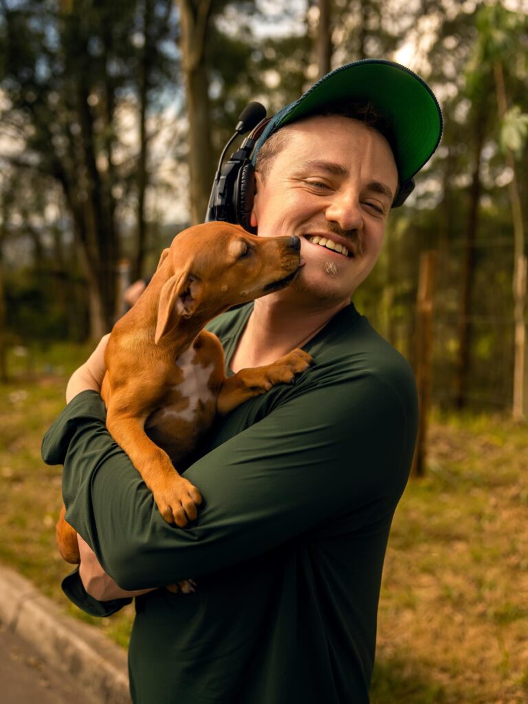 A imagem mostra o diretor do filme sorridente segurando um filhote de cachorro caramelo nos braços. O animal lambe o rosto dele com carinho, enquanto estão ao ar livre, em um ambiente com árvores e luz natural. A cena transmite afeto e alegria.