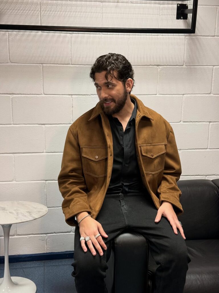 A imagem mostra Gustavo Mioto sentado em um sofá preto, vestindo jaqueta marrom, camisa preta e calça preta. Ele olha para o lado com expressão descontraída e usa vários anéis prateados nas mãos. Ao lado, há uma mesinha branca redonda.