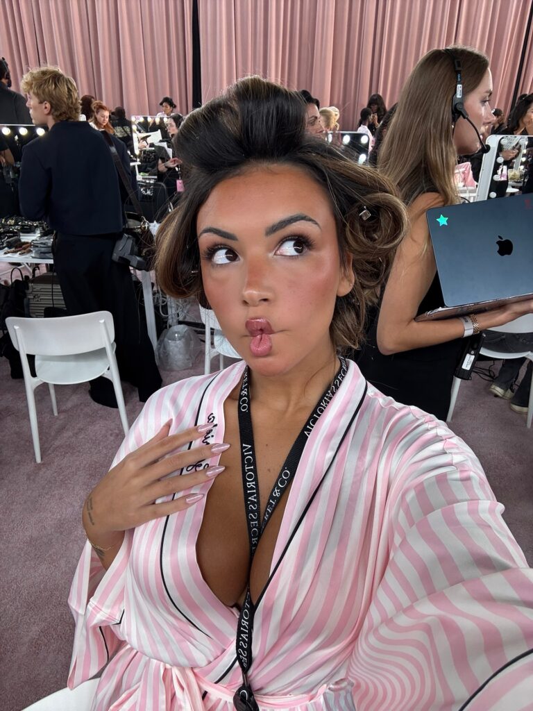 A imagem mostra uma selfie da influencer Gabi Moura está fazendo um "biquinho de peixe", com uma expressão facial divertida e a pose sugerindo um momento de descontração e brincadeira. Ela tem pele bronzeada e cabelo castanho escuro com rolos de cabelo, sugerindo que está se preparando para um evento. Seu rosto é lindamente maquiado, com blush nas bochechas e sombra nos olhos. Ela está usando um roupão rosa listrado com detalhes em branco e preto, e um cordão de pescoço com o logotipo "VICTORIA'S SECRET & CO".