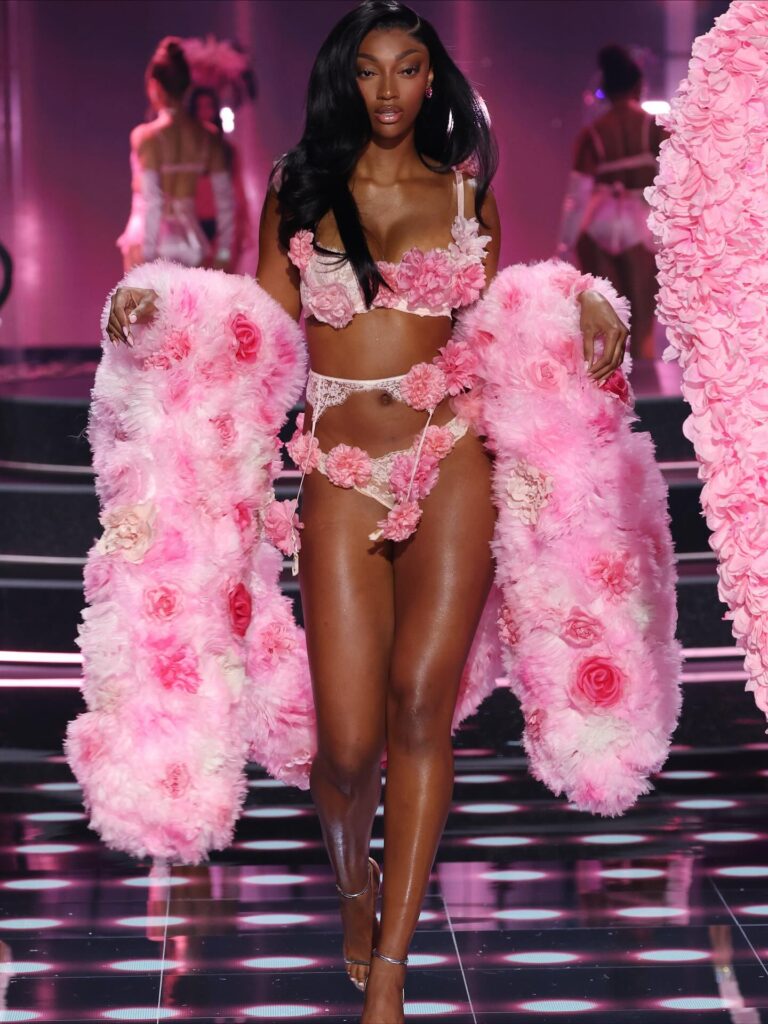 A imagem mostra a modelo de pele preta e atleta Angel Reese desfilando. Ela  caminha pela passarela, exibindo um conjunto de lingerie delicado e uma estola exuberante em tons de rosa. 
