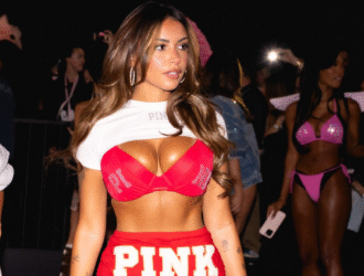 A imagem mostra a influencer Gabi Moura desfilando em um evento de moda, vestindo um conjunto vermelho com a palavra “PINK” em destaque. Ela usa um top e uma saia curtos, com cabelo solto e maquiagem marcada. Ao fundo, outras modelos aparecem com looks em tons de rosa e preto, enquanto o público registra o momento com celulares. O ambiente é iluminado e vibrante, típico de um desfile