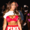 A imagem mostra a influencer Gabi Moura desfilando em um evento de moda, vestindo um conjunto vermelho com a palavra “PINK” em destaque. Ela usa um top e uma saia curtos, com cabelo solto e maquiagem marcada. Ao fundo, outras modelos aparecem com looks em tons de rosa e preto, enquanto o público registra o momento com celulares. O ambiente é iluminado e vibrante, típico de um desfile