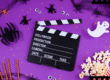 A imagem mostra um cenário com fundo roxo, com detalhes típicos do Halloween, como teias de areia no canto, morceguinhos pretos, olhos de medo e fantasma. No canto direito inferior está um balde de pipoca, e no centro há um símbolo de gravação de cenas em filmes