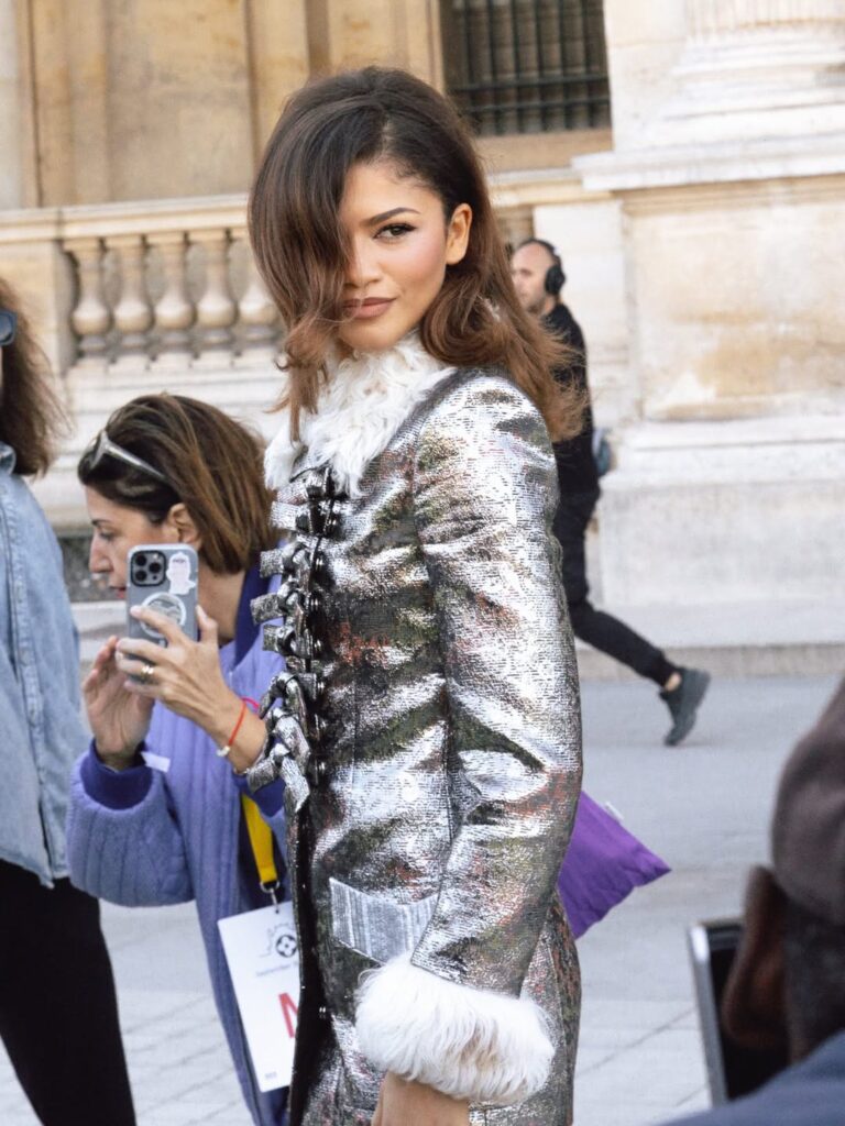 A imagem mostra a atriz Zendaya usando um elegante casaco  longo metalizado prateado com detalhes de pelúcia branca nos punhos e gola. O look sofisticado e futurista destaca sua presença marcante, enquanto ela posa com confiança diante das câmeras.