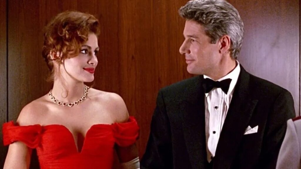 A imagem mostra uma cena do filme "Uma Linda Mulher (1990)". Nela, Julia Roberts está à esquerda, vestindo um elegante vestido vermelho decotado com ombros à mostra e luvas brancas longas. Ela usa um colar chamativo e seu cabelo ruivo está preso em um penteado volumoso e ondulado. À direita, Richard Gere aparece em um smoking preto clássico, com camisa branca e gravata borboleta preta. Ambos estão se olhando com um sorriso sutil, transmitindo uma atmosfera de charme e conexão entre os personagens. O fundo é de madeira clara, sugerindo que estão em um ambiente interno sofisticado, possivelmente um hotel. 