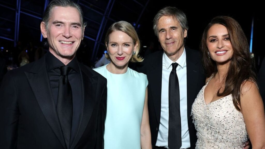 A imagem mostra os atores Billy Crudup, Naomi Watts, o cineasta Walter Salles e a atriz Penélope Cruz posando juntos em um evento de gala. Todos estão elegantemente vestidos: Crudup usa um terno preto com gravata da mesma cor, Naomi Watts veste um vestido azul-claro e segura uma clutch prateada, Walter Salles aparece de terno escuro com camisa branca e gravata preta, e Penélope Cruz usa um vestido bege com aplicações brilhantes. O grupo sorri para a câmera em um ambiente sofisticado e bem iluminado, típico de cerimônias de premiação ou eventos cinematográficos.