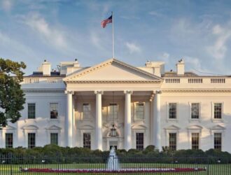 A imagem mostra a Casa Branca, residência oficial e local de trabalho do Presidente dos Estados Unidos. É um edifício grande, de estilo neoclássico, pintado de branco. Na frente, há um pórtico com colunas altas e um frontão triangular no topo. No alto do prédio, está hasteada uma bandeira dos Estados Unidos. O edifício é cercado por um gramado bem cuidado e uma cerca preta de ferro. O céu está claro, destacando ainda mais a imponência da construção