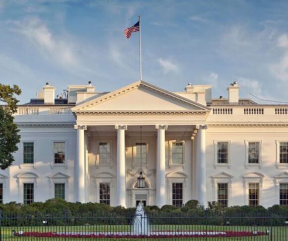 A imagem mostra a Casa Branca, residência oficial e local de trabalho do Presidente dos Estados Unidos. É um edifício grande, de estilo neoclássico, pintado de branco. Na frente, há um pórtico com colunas altas e um frontão triangular no topo. No alto do prédio, está hasteada uma bandeira dos Estados Unidos. O edifício é cercado por um gramado bem cuidado e uma cerca preta de ferro. O céu está claro, destacando ainda mais a imponência da construção