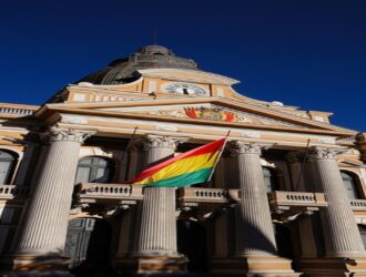 A imagem mostra o Palácio Legislativo, localizado em La Paz, capital da Bolívia. Este edifício histórico é a sede da Assembleia Legislativa Plurinacional do país. Sua arquitetura é de estilo neoclássico, com colunas altas, janelas em arco e uma cúpula imponente no topo. Acima da entrada principal, há um relógio e o brasão de armas da Bolívia. Em frente ao prédio, a bandeira nacional boliviana está hasteada com destaque. O céu está limpo e azul intenso, o que realça ainda mais a beleza e imponência da construção