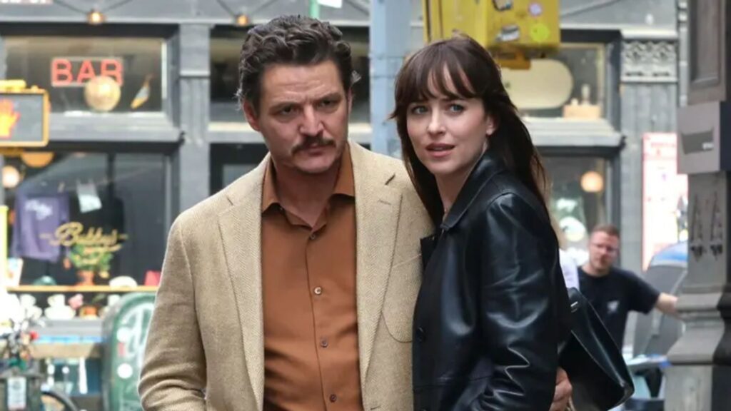 Na imagem, os atores Pedro Pascal e Dakota Johnson aparecem caminhando juntos em uma rua movimentada, em cena do filme Amores Materialistas.Pedro Pascal veste um paletó bege e camisa marrom, com expressão séria e o braço envolto na cintura de Dakota Johnson, que usa uma jaqueta de couro preta e olha ligeiramente para o lado, com semblante atento. O fundo urbano, com fachadas de bares e letreiros, reforça o clima contemporâneo e realista da trama.