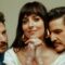 Na imagem, aparecem os atores Chris Evans, Dakota Johnson e Pedro Pascal, que estrelam o filme "Amores Materialistas". Dakota Johnson está ao centro, apoiando os cotovelos sobre uma mesa, com as mãos entrelaçadas e um leve sorriso. À esquerda, Chris Evans a observa com expressão suave e corpo inclinado em sua direção. À direita, Pedro Pascal se aproxima dela, com o rosto próximo ao dela e olhar contemplativo. A cena transmite intimidade e ternura, representando o triângulo amoroso central do longa