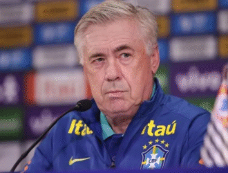 Técnico da Seleção Brasileira, Ancelotti, com cabelos grisalhos, vestindo um agasalho azul da Seleção com o escudo da CBF e o logotipo da patrocinadora Itaú. Ele está em uma coletiva de imprensa, sentado diante de um fundo com logos de marcas e olhando seriamente para a frente
