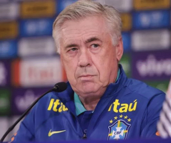Técnico da Seleção Brasileira, Ancelotti, com cabelos grisalhos, vestindo um agasalho azul da Seleção com o escudo da CBF e o logotipo da patrocinadora Itaú. Ele está em uma coletiva de imprensa, sentado diante de um fundo com logos de marcas e olhando seriamente para a frente