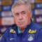 Técnico da Seleção Brasileira, Ancelotti, com cabelos grisalhos, vestindo um agasalho azul da Seleção com o escudo da CBF e o logotipo da patrocinadora Itaú. Ele está em uma coletiva de imprensa, sentado diante de um fundo com logos de marcas e olhando seriamente para a frente