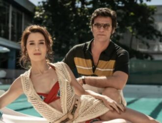 Cena da série "Ângela Diniz: Assassinada e Condenada", da HBO Max. À beira de uma piscina, Marjorie Estiano, que interpreta Ângela Diniz, aparece reclinada em uma espreguiçadeira, usando maiô vermelho e colete de crochê claro, com expressão serena. Atrás dela, Emílio Dantas, no papel de Doca Street, usa óculos escuros e camisa de listras amarelas e pretas, observando-a com postura confiante. Ao fundo, um prédio moderno e guarda-sóis compõem o cenário de luxo típico dos anos 1970