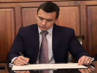 A imagem mostra Daniel Noboa, presidente do Equador, vestindo um terno escuro, camisa clara e gravata, sentado à frente de uma mesa de madeira polida. Ele está escrevendo ou assinando um documento com uma caneta, demonstrando foco e seriedade.