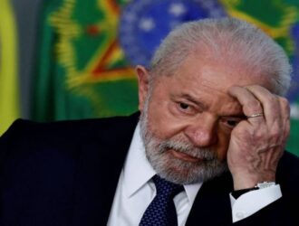 O presidente Luiz Inácio Lula da Silva aparece sentado, com expressão pensativa. Ele tem pele clara, cabelos brancos e barba branca. Veste um terno escuro, camisa branca e gravata preta. Está com o cotovelo apoiado na mesa e a mão esquerda encostada no rosto, como se estivesse refletindo ou preocupado