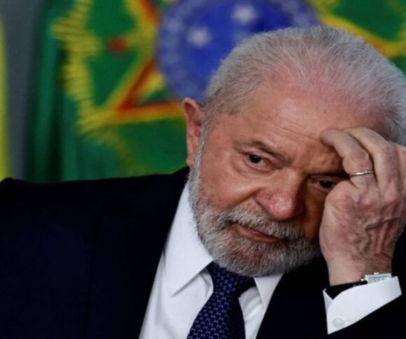 O presidente Luiz Inácio Lula da Silva aparece sentado, com expressão pensativa. Ele tem pele clara, cabelos brancos e barba branca. Veste um terno escuro, camisa branca e gravata preta. Está com o cotovelo apoiado na mesa e a mão esquerda encostada no rosto, como se estivesse refletindo ou preocupado