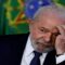 O presidente Luiz Inácio Lula da Silva aparece sentado, com expressão pensativa. Ele tem pele clara, cabelos brancos e barba branca. Veste um terno escuro, camisa branca e gravata preta. Está com o cotovelo apoiado na mesa e a mão esquerda encostada no rosto, como se estivesse refletindo ou preocupado