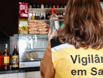 Mulher com blusa estampada com a palavra 'Vigilância Sanitária', analisando uma garrafa de bebida alcoólica