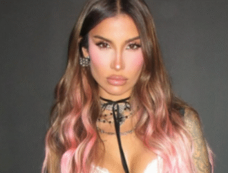 Bianca Andrade posa em frente a um fundo preto, exibindo o novo visual com cabelos longos em tom castanho nas raízes e rosa nas pontas. Ela usa uma blusa preta justa com alças finas e detalhe branco de renda, calça jeans preta e acessórios prateados, incluindo colares e brincos. Suas tatuagens nos braços estão visíveis, e a expressão é séria e confiante, destacando o estilo moderno e ousado da empresária