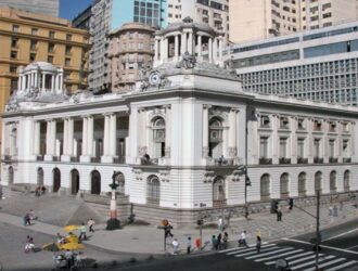A imagem mostra a Câmara Municipal do Rio de Janeiro, local onde será votada a Lei Anti-Oruam, um edifício histórico e imponente localizado em uma esquina movimentada da cidade. Sua arquitetura é de estilo clássico, com colunas, detalhes ornamentais e estátuas no topo, transmitindo uma atmosfera de solenidade e tradição