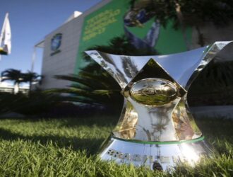 A imagem mostra o troféu do Campeonato Brasileiro Série A, principal competição de futebol do Brasil. O troféu está posicionado sobre uma área gramada bem cuidada, em frente a um prédio moderno