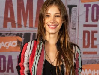 A imagem mostra a atriz Caroline Dallarosa sorrindo em frente a um painel com as palavras “Malhação” e “Toda Forma de Amar”. Ela tem cabelos longos e castanhos, veste uma jaqueta colorida com listras vermelhas, pretas e verdes sobre uma blusa preta e usa um colar delicado. O fundo apresenta tons de laranja, vermelho e cinza, remetendo à identidade visual da novela juvenil da TV Globo.