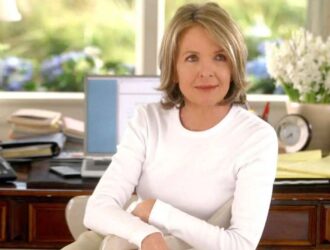 A atriz Diane Keaton sentada em um ambiente iluminado, usando uma blusa branca de mangas compridas. Ela está com expressão serena, levemente sorridente, e tem cabelos loiros curtos. Ao fundo, há uma mesa com livros empilhados, um computador, um relógio e um vaso de flores brancas, próximos a uma janela ampla por onde entra luz natural