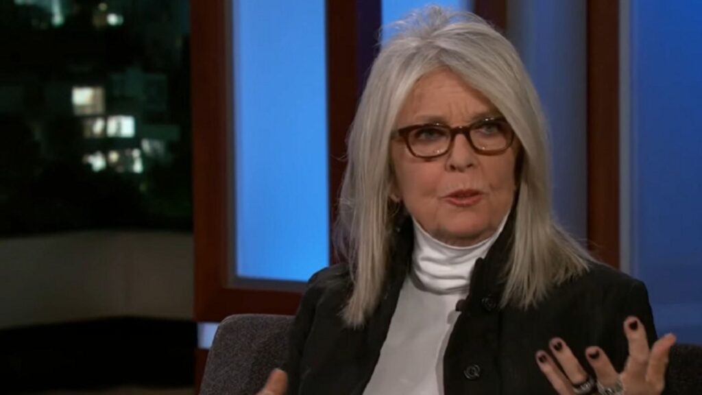 Diane Keaton está sentada em um estúdio de televisão, vestindo uma blusa branca de gola alta e um casaco preto. Ela usa óculos de armação escura e esmalte preto nas unhas, gesticulando com uma das mãos enquanto fala. O fundo mostra janelas com luzes de cidade ao anoitecer e uma parede azul iluminada pelo cenário do programa.