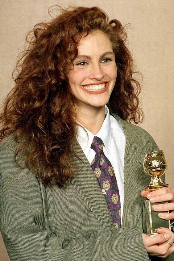 Julia Roberts, ícone dos cabelos ondulados, aparece com cabelo ruivo e longo estilizado com um topete. Ela usa um terno oversized verde, uma camisa de botões branca e gravata de fundo verde escuro com estampa de flores roxas e amarelas enquanto sorri e segura um troféu.