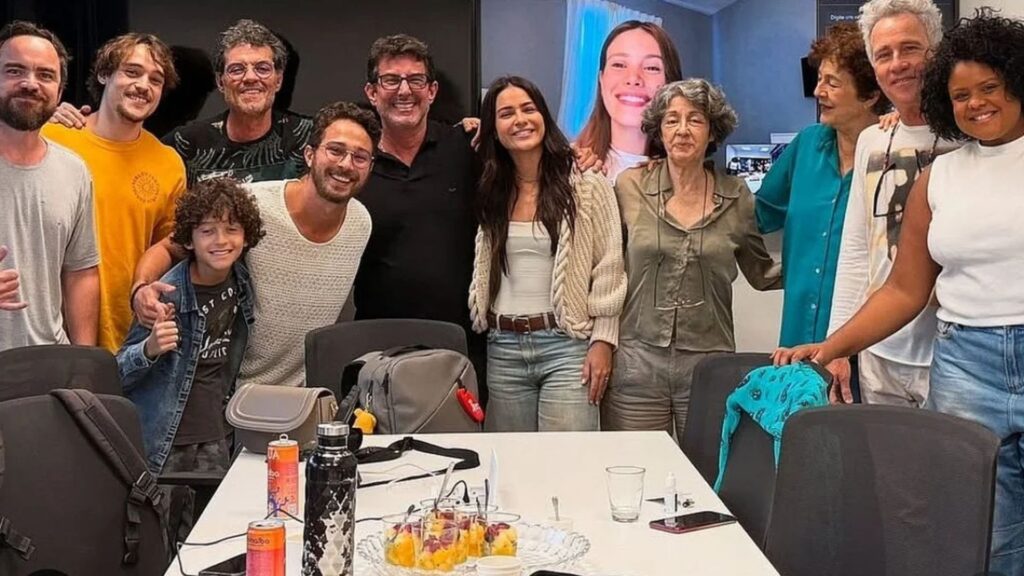 Bastidores da produção da série "Vidas Paralelas". Os atores e atrizes posam sorrindo em torno de uma mesa de reunião, em clima descontraído. Ao fundo, é possível ver uma atriz participando por videoconferência. Sobre a mesa, itens pessoais, copos e petiscos. A foto transmite o avanço das gravações e o ambiente colaborativo da produção