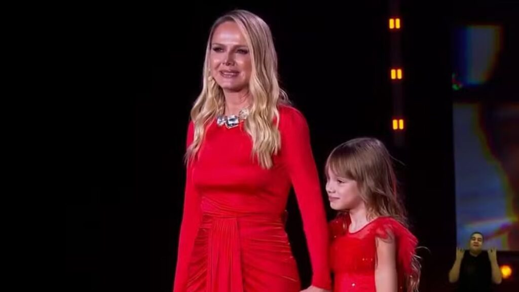 A imagem mostra Eliana e uma criança em um palco, durante o "Upfront Globo 2026". À esquerda, Eliana está de cabelos longos e loiros, veste um vestido vermelho elegante com mangas longas e detalhes franzidos na cintura. Ela segura a mão de uma menina à sua direita, que também está vestida de vermelho, em um vestido com detalhes de tule nas mangas. Ambas olham para o público, com expressões serenas e discretamente sorridentes. No canto inferior direito, há uma intérprete de libras em fundo escuro. O fundo do palco é predominantemente preto, com algumas luzes verticais amarelas iluminando a cena.