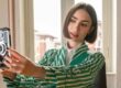 Lilly Collins está com cabelo curto e liso, vestindo um blazer listrado em verde e branco sobre uma camisa branca, segura o celular em posição de selfie. Ela está em um ambiente interno iluminado pela luz natural que entra por uma janela, com detalhes decorativos nas paredes e prédios visíveis ao fundo