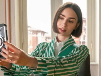 Lilly Collins está com cabelo curto e liso, vestindo um blazer listrado em verde e branco sobre uma camisa branca, segura o celular em posição de selfie. Ela está em um ambiente interno iluminado pela luz natural que entra por uma janela, com detalhes decorativos nas paredes e prédios visíveis ao fundo
