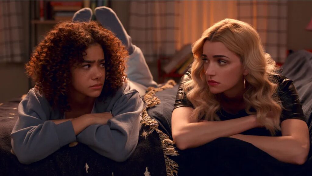Na imagem, estão as personagens Ginny Miller e Georgia Miller, interpretadas por Antonia Gentry e Brianne Howey, respectivamente, na série "Ginny & Georgia". As duas aparecem deitadas de bruços sobre uma cama, encarando-se com expressões sérias e reflexivas. Ginny veste um moletom azul-claro, enquanto Georgia usa uma blusa preta e exibe cabelos loiros ondulados.A cena transmite um momento de tensão e diálogo íntimo entre mãe e filha, destacando a complexidade emocional que marca a relação das protagonistas.