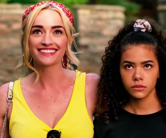 Na imagem, aparecem as personagens Georgia Miller, interpretada por Brianne Howey, e Ginny Miller, vivida por Antonia Gentry, da série "Ginny & Georgia". Georgia está sorridente, vestindo uma blusa amarela e uma faixa vermelha no cabelo, com o braço sobre o ombro da filha. Ao lado, Ginny exibe uma expressão séria e pensativa, usando uma blusa escura e um penteado com scrunchie. A cena simboliza o contraste entre a personalidade expansiva da mãe e a introspecção da filha, um dos eixos centrais da trama.