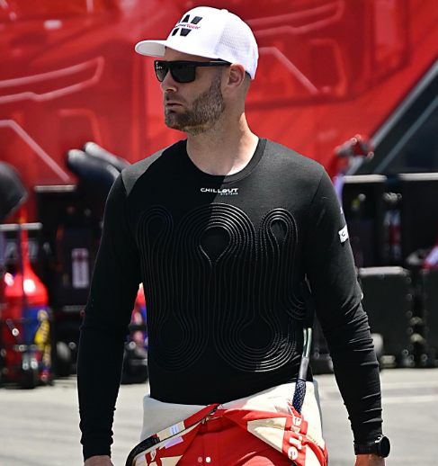 O piloto Shane van Gisbergen aparece em pé, vestindo um boné branco, óculos escuros e o colete de resfriamento preto da marca Chillout, que possui tubos internos para circulação de líquido refrigerante. Ele está com parte do macacão de corrida vermelho e branco abaixado até a cintura, em um ambiente de paddock com equipamentos de box ao fundo.