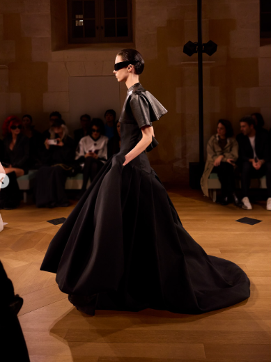 Modelo desfilando em passarela com um vestido preto volumoso de alta costura, com estrutura dramática e mangas curtas rígidas, usando óculos escuros e cabelos molhados, em ambiente elegante com público assistindo ao fundo.
