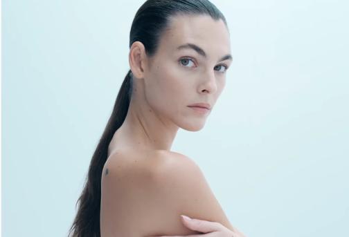 Mulher de cabelos castanhos escuros presos em um rabo de cavalo com efeito molhado, olhando sobre o ombro em direção à câmera. Ela está com os ombros nus e posando contra um fundo branco suave, transmitindo uma estética minimalista e clean.