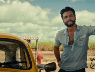 Wagner Moura posa com camisa azul claro de botões, com um braço apoiado em uma parede e uma mão na cintura, enquanto ao lado de um carro amarelo