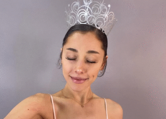 Ariana Grande aparece em frente a um fundo de cor neutra, com os olhos fechados e sorrindo levemente. Ela a coroa que sua personagem Glinda tem como acessório no filme Wicked.