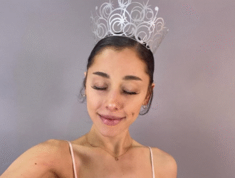 Ariana Grande aparece em frente a um fundo de cor neutra, com os olhos fechados e sorrindo levemente. Ela a coroa que sua personagem Glinda tem como acessório no filme "Wicked"