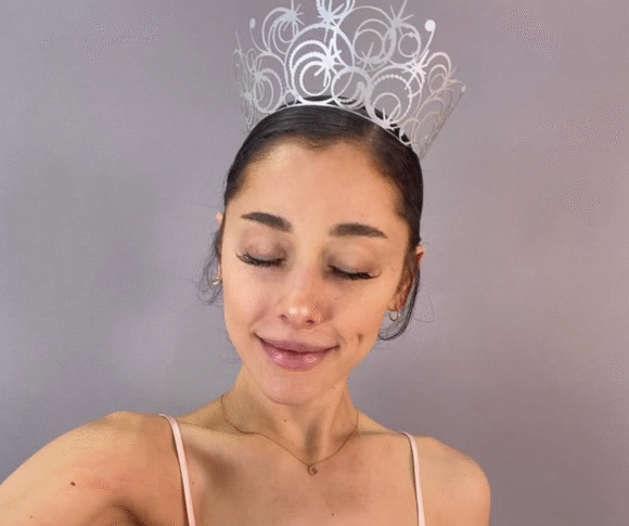 Ariana Grande aparece em frente a um fundo de cor neutra, com os olhos fechados e sorrindo levemente. Ela a coroa que sua personagem Glinda tem como acessório no filme Wicked.